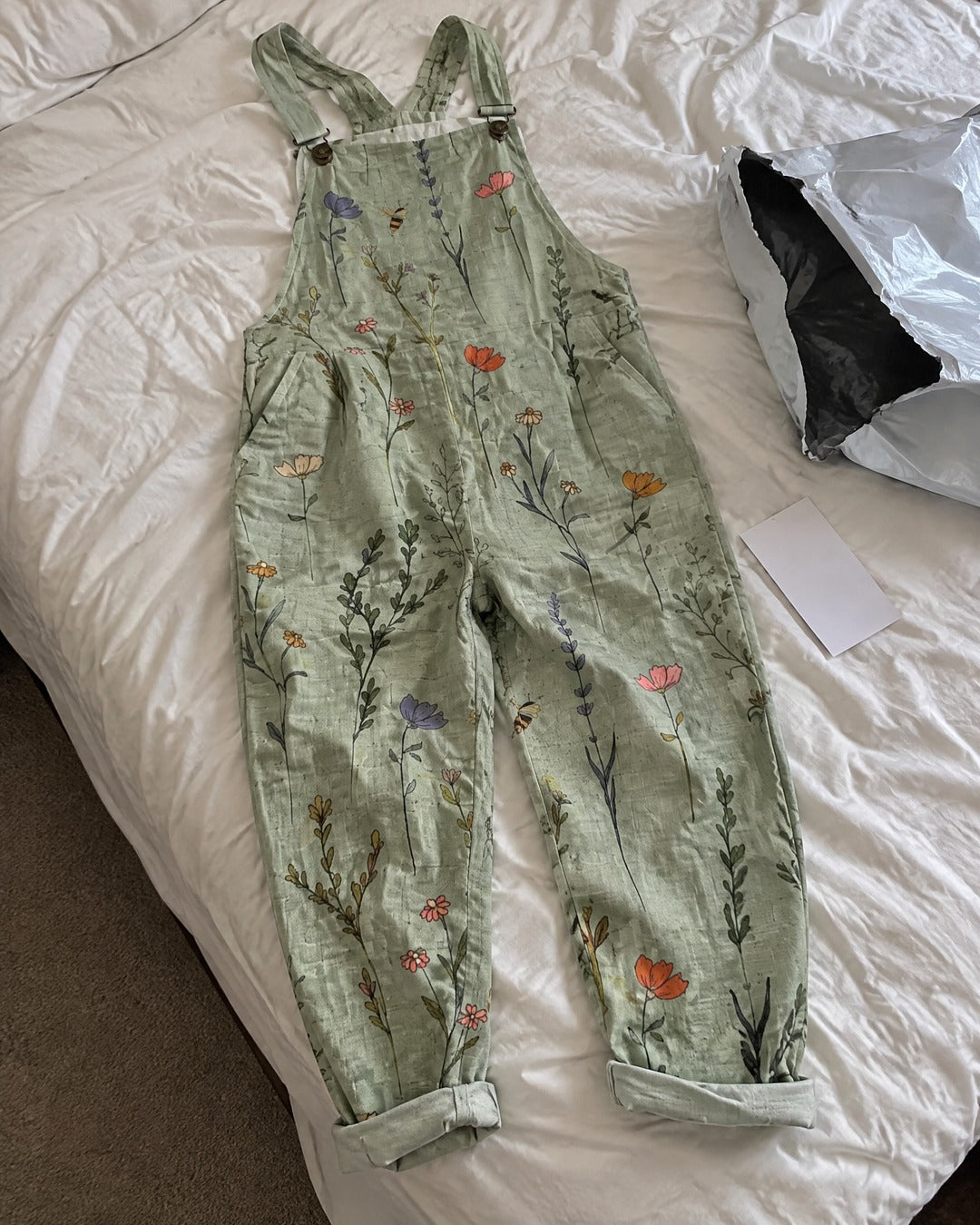 Sage green floral printed overalls laid on bed showing adjustable shoulder straps wide legs and flower print