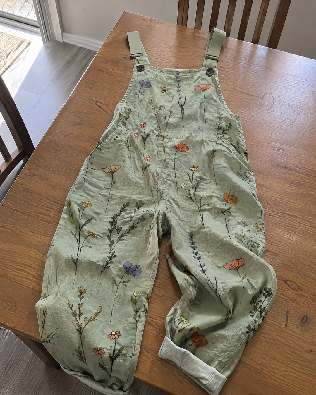Sage green botanical print overalls laid on wooden dining table with adjustable straps and roomy legs