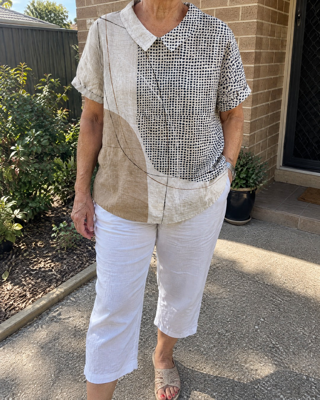 Mature woman wearing Lana abstract dots and lines shirt with white cropped trousers outside home
